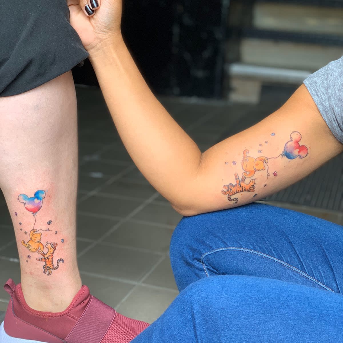 Matching tattoos of Winnie the Pooh and Tigger with colorful Mickey Mouse-shaped balloons on two people's arms and legs